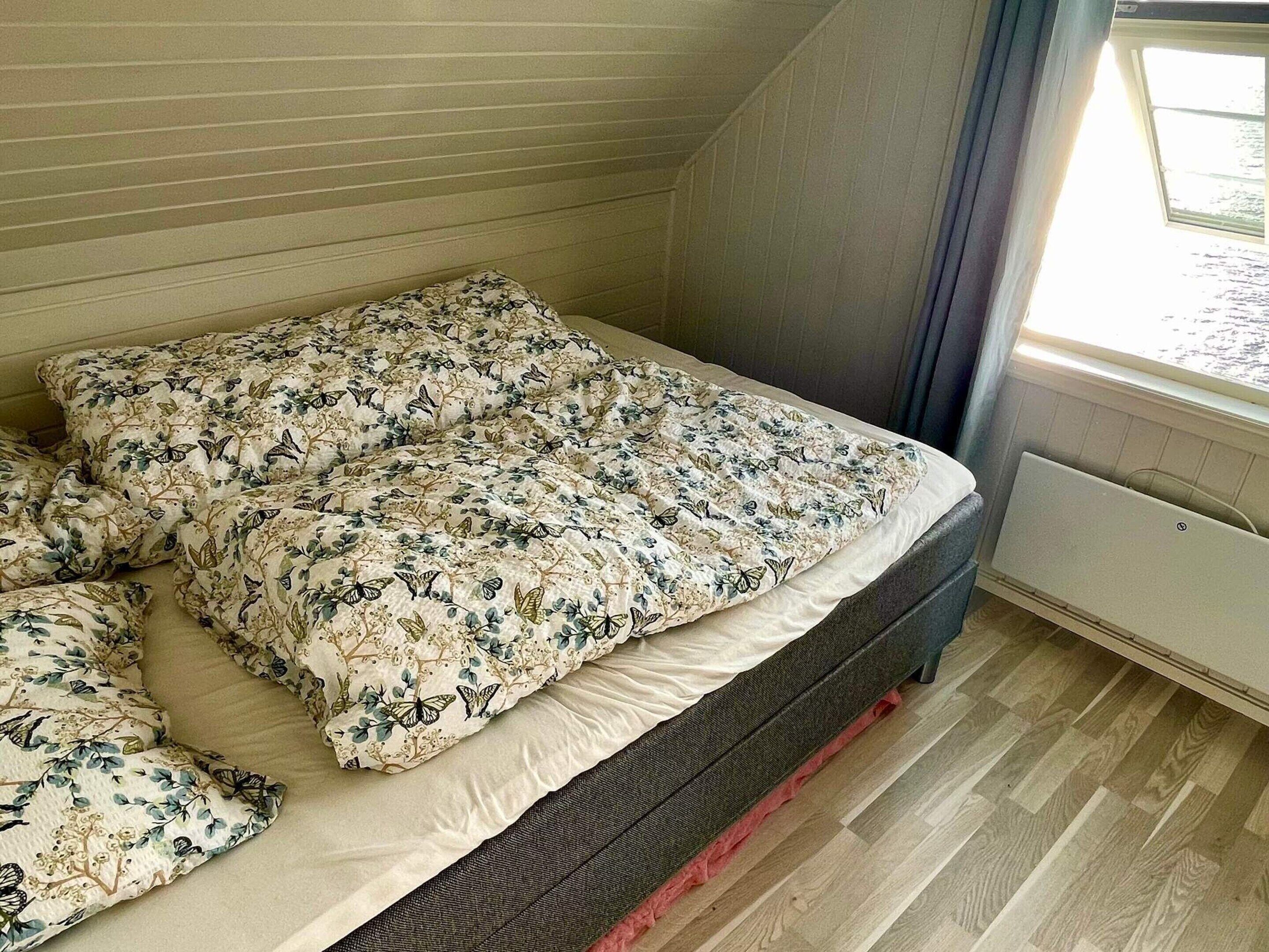 A bedroom with a bed covered in floral-patterned bedding, a window with blinds, and a radiator on the floor.