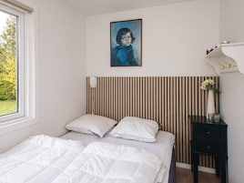 A bedroom with a bed, pillows, a nightstand, a vase with flowers, a painting, and a window.