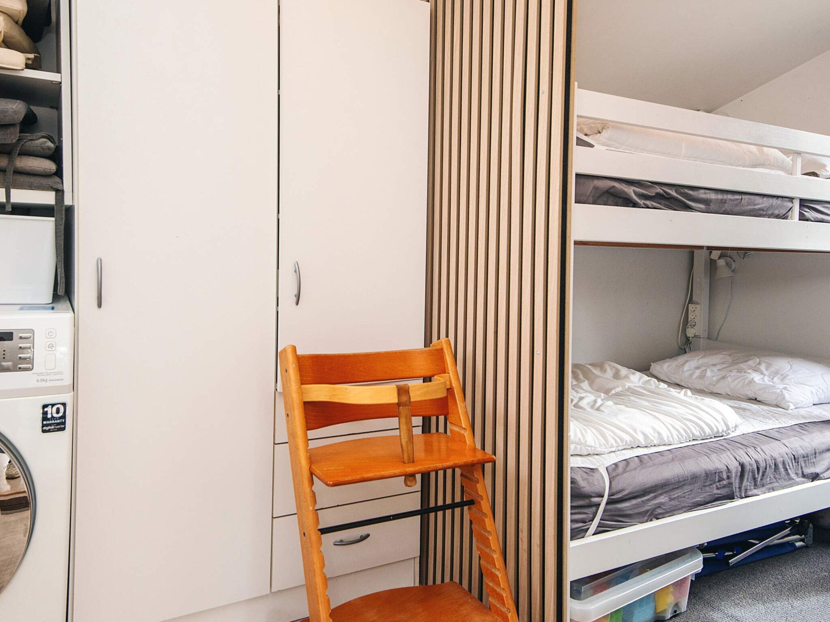Una cama litera con cabecera blanca, una silla de madera y un armario blanco con panel de control.