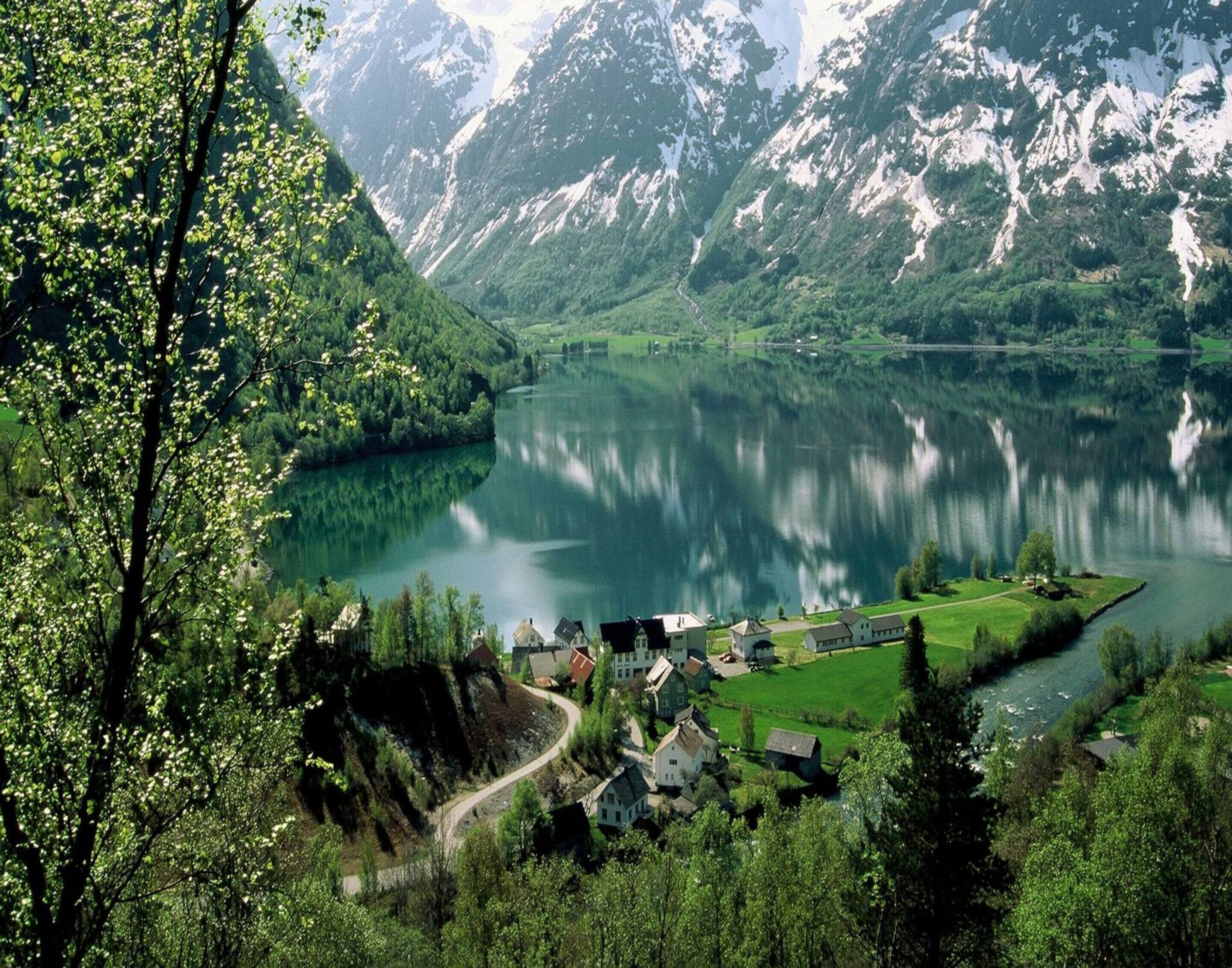 A lakeside village with traditional houses, a winding road, and mountains in the background.