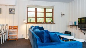 A room with a blue sofa, a small table, a TV, and a window with a view of greenery.