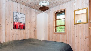 A room with wooden walls, a bed with a dark quilt, a window with a view of greenery, a framed picture on the wall, and a door.