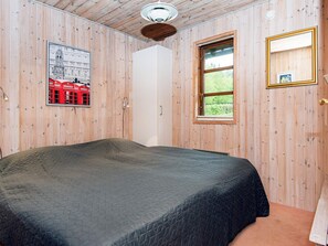 A room with wooden walls, a bed with a dark quilt, a window with a view of greenery, a framed picture on the wall, and a door. - 4 Person Holiday Home in Toftlund (Toftlund)