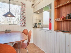 A kitchen with a dining table, chairs, a hanging light fixture, a window with curtains, and a wooden shelf with kitchen items. - 4 Person Holiday Home in Aakirkeby (Akirkeby)