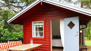 A red cabin with a wooden table and striped cushions, a window, and a door leading to a bedroom. - 6 Person Holiday Home in Monsteras (Monsteras)