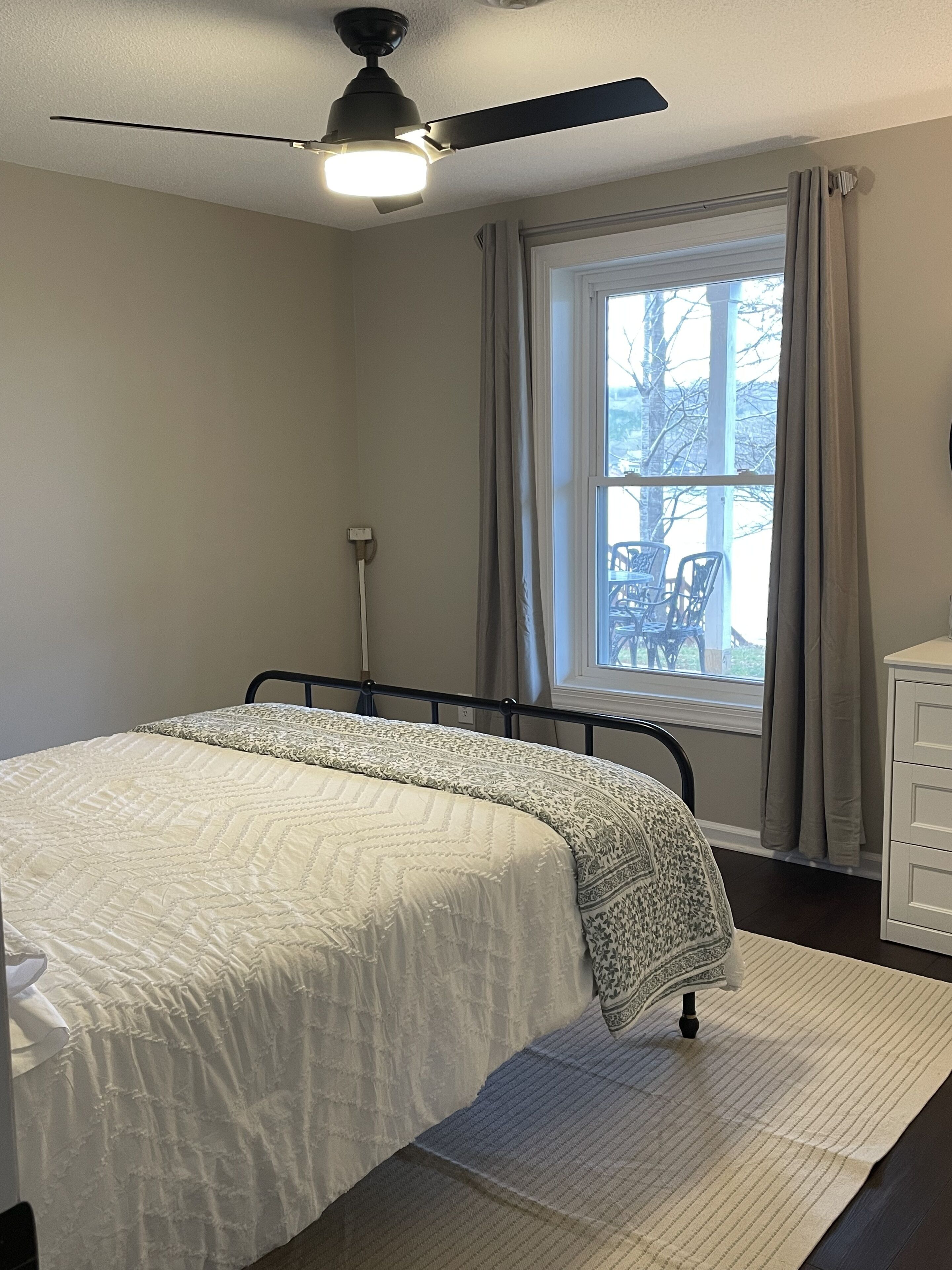 Un dormitorio con cama, ventilador de techo, ventana con cortinas, una silla afuera y cómoda.