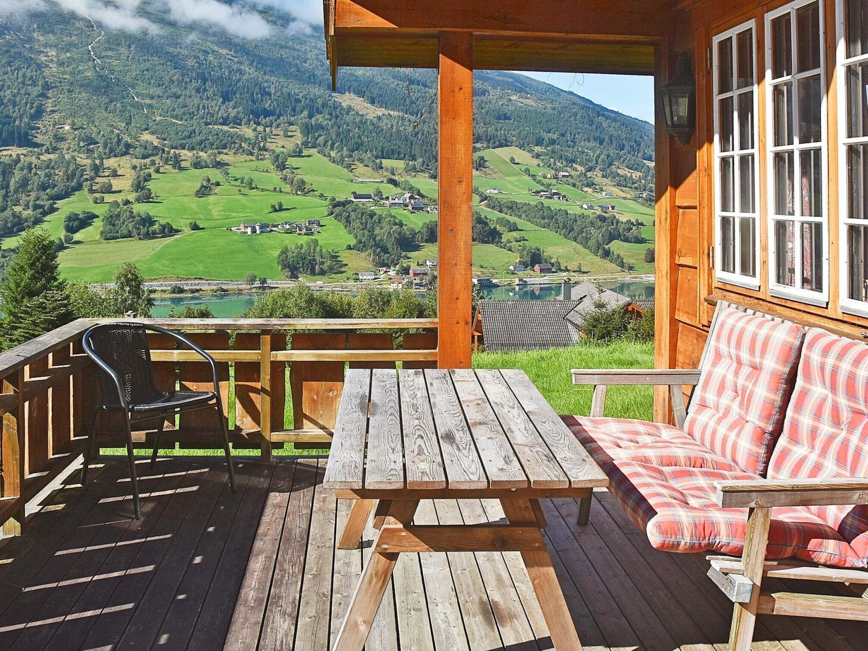 A wooden deck with a table and chairs, overlooking a scenic valley with houses and a lake.