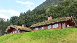 Une cabane en bois, dotée d’un toit en pente, située sur une colline herbeuse, avec une montagne boisée en arrière-plan.