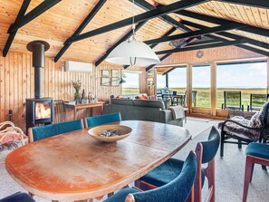 A cozy interior with a wooden ceiling, a fireplace, a dining table, and a view through large windows.