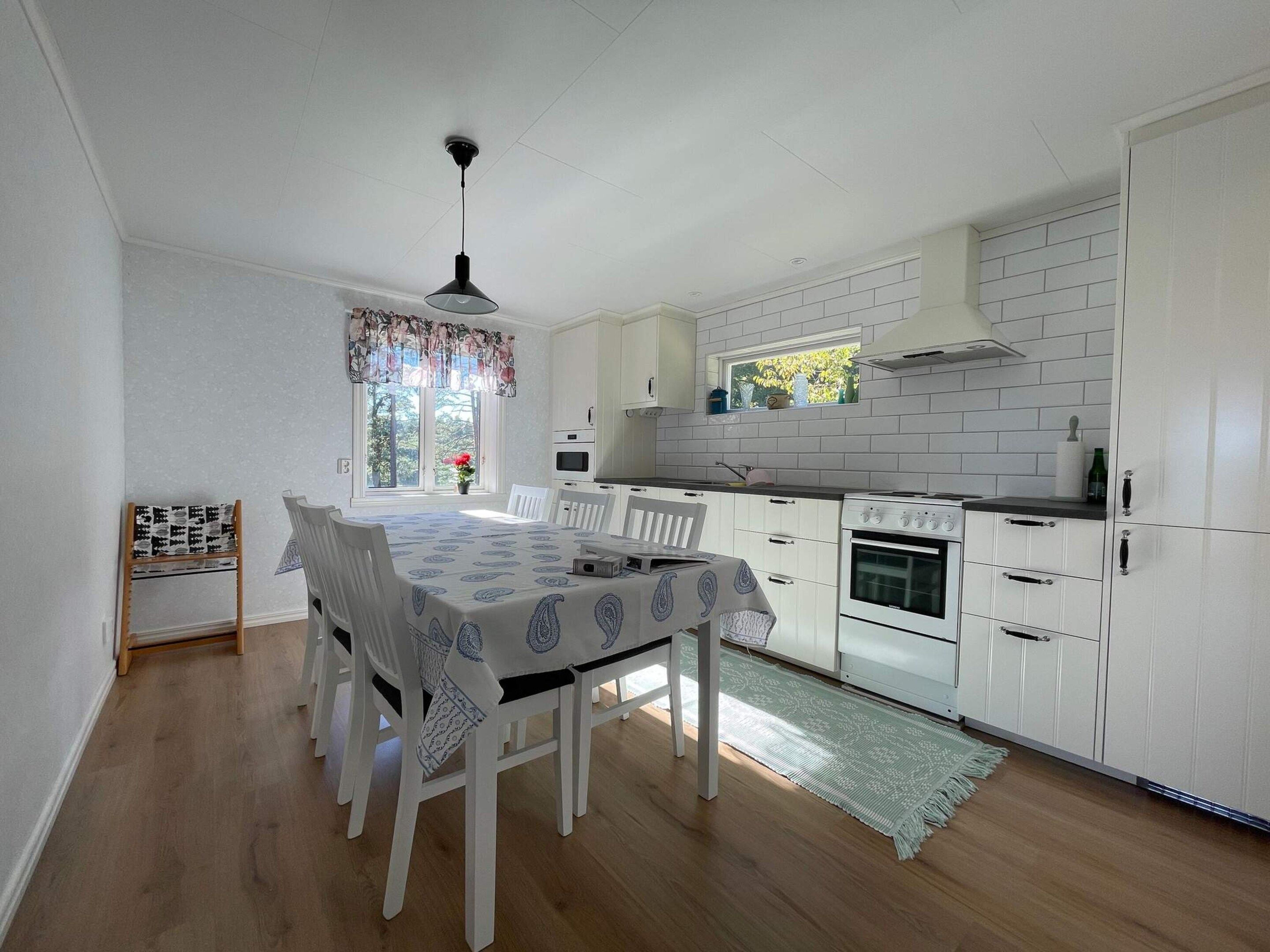 Une cuisine avec des meubles blancs, une table à manger avec des chaises, un luminaire suspendu et une fenêtre donnant sur des arbres.