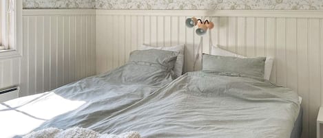 Una cama individual con cabecera blanca y dos almohadas, un florero verde en una repisa arriba y una ventana a la izquierda.