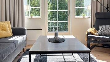 A living room with a grey sofa, a black coffee table, and a grey armchair. There are two windows with curtains, a lamp, and a small table with a candle and a decorative item.