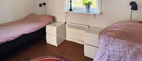 A bedroom with two beds, a bedside table with a lamp, a window with a plant, and a radiator.