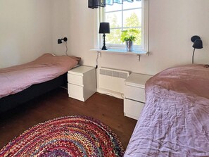A bedroom with two beds, a bedside table with a lamp, a window with a plant, and a radiator.