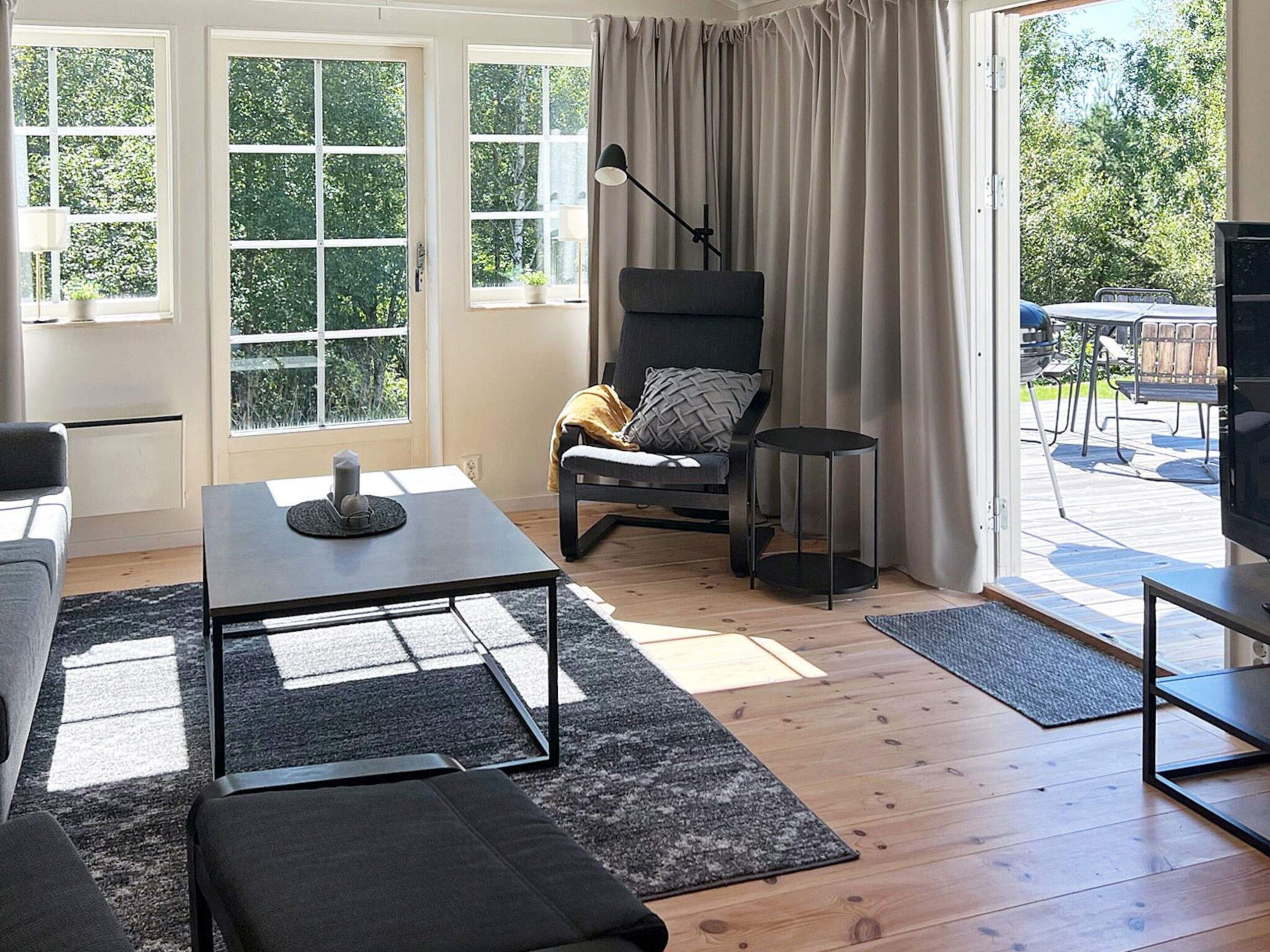 A modern living room with a sofa, a coffee table, a chair, and a television. There are large windows with curtains, a door, and a view of the outdoors.