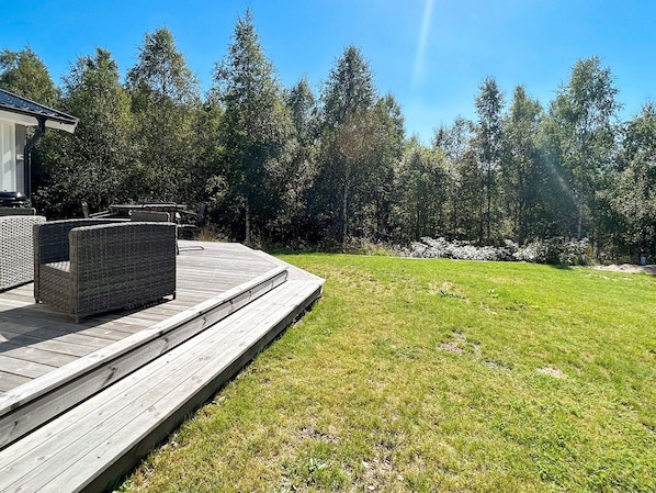 A wooden deck with outdoor furniture, surrounded by a grassy lawn and dense trees.