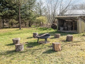 A rustic outdoor setting with a metal fire pit surrounded by tree stumps, a wooden shed with logs, and a forested background. - 5 Person Holiday Home in Tarm (Tarm)