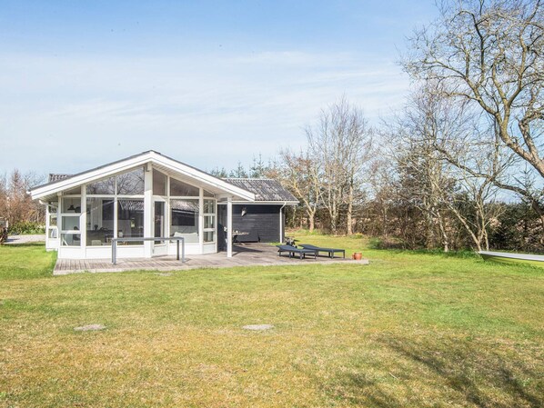 Una casa moderna con un amplio ventanal de vidrio, un patio con mobiliario exterior y un área verde circundante.