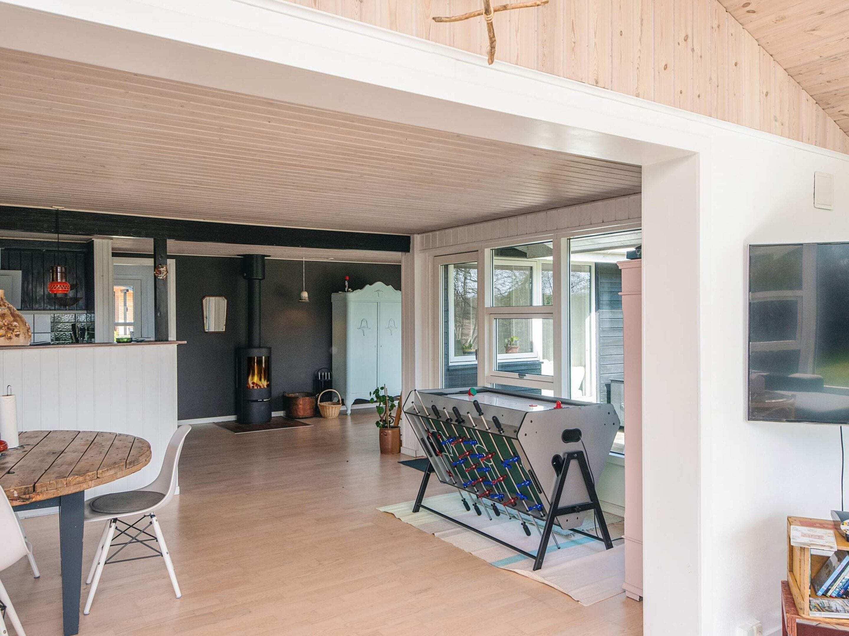 Un interior moderno con techo de madera, una chimenea central, una zona de comedor con mesa y sillas, y una mesa de futbolín.