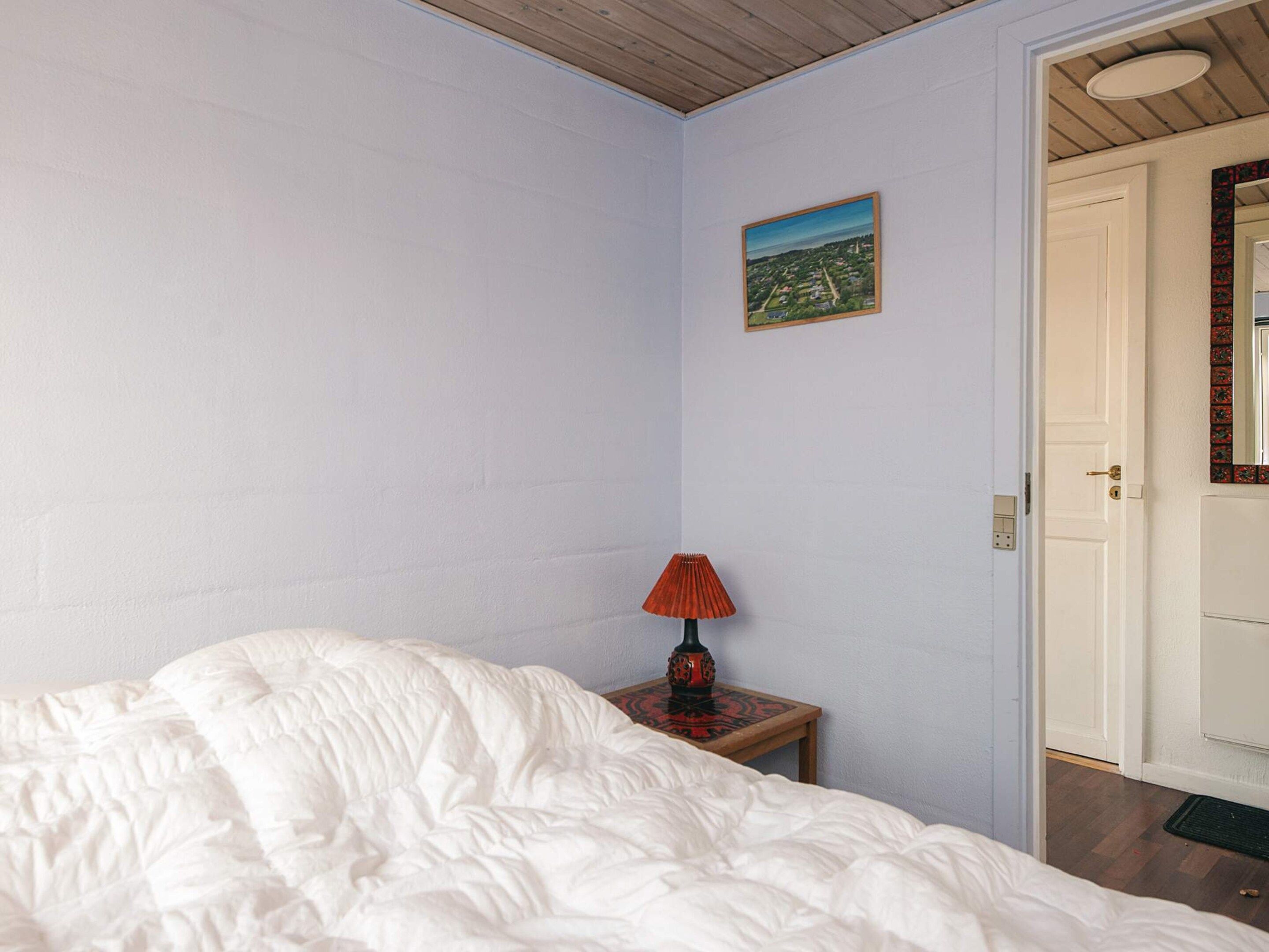 A bedroom with a bed, a nightstand with a lamp, a door, and a wall-mounted picture.