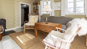 Un salon chaleureux avec une table basse en bois, un fauteuil en osier, un fauteuil moelleux blanc et un canapé gris. On y trouve une bibliothèque remplie de livres et une carte accrochée au mur, ainsi qu’une fenêtre avec des rideaux.
