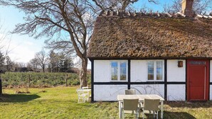 Une maison traditionnelle à toit de chaume, avec une façade blanche et des éléments décoratifs noirs, une porte rouge et une petite table entourée de chaises dans le jardin.