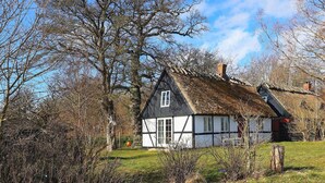 Une maison traditionnelle à colombages, dotée d’un toit à forte pente, entourée d’arbres et bordée d’un chemin de terre.