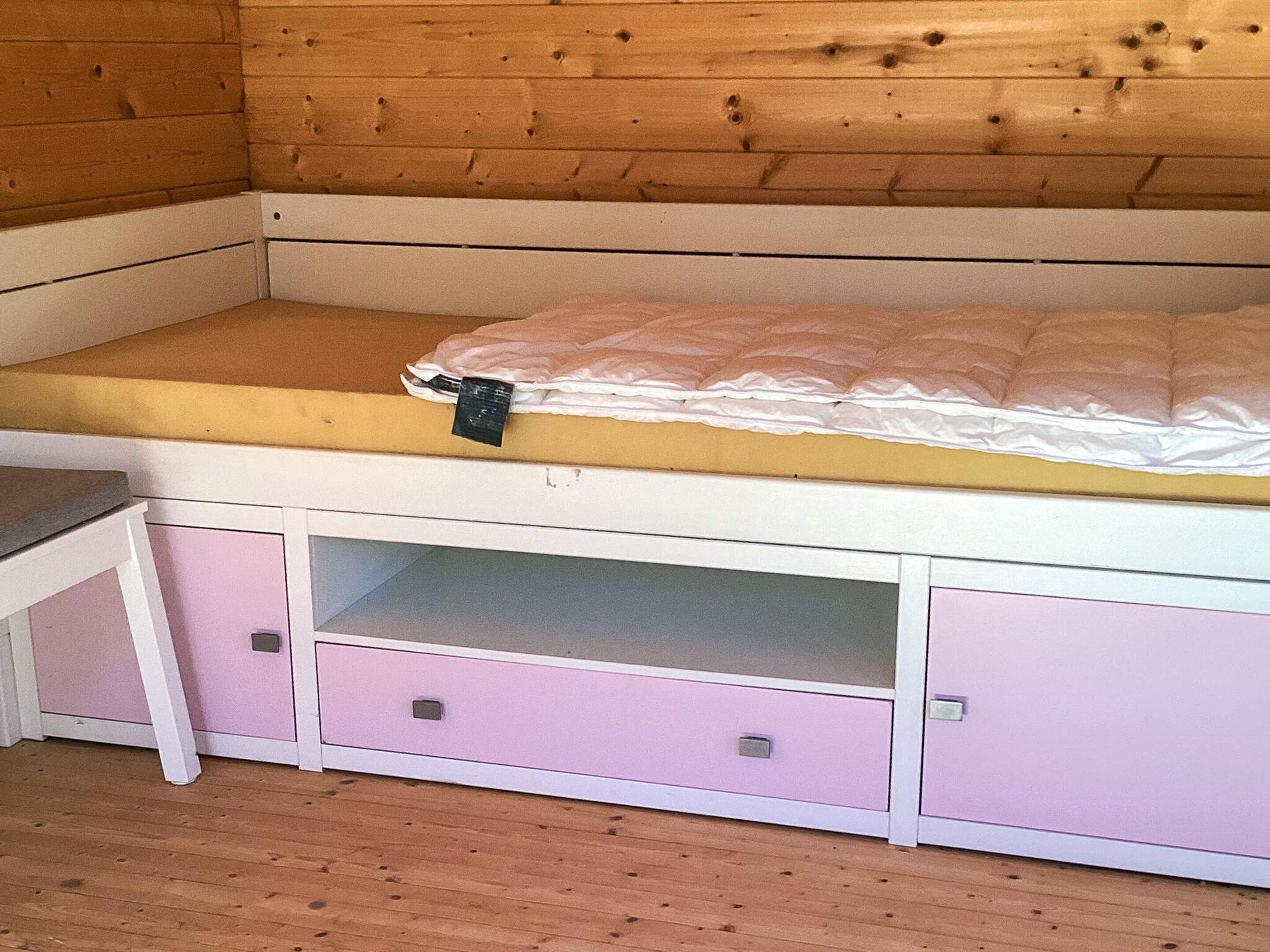 A corner bedroom with a wooden ceiling, a single bed with a white mattress, and a small desk with drawers.