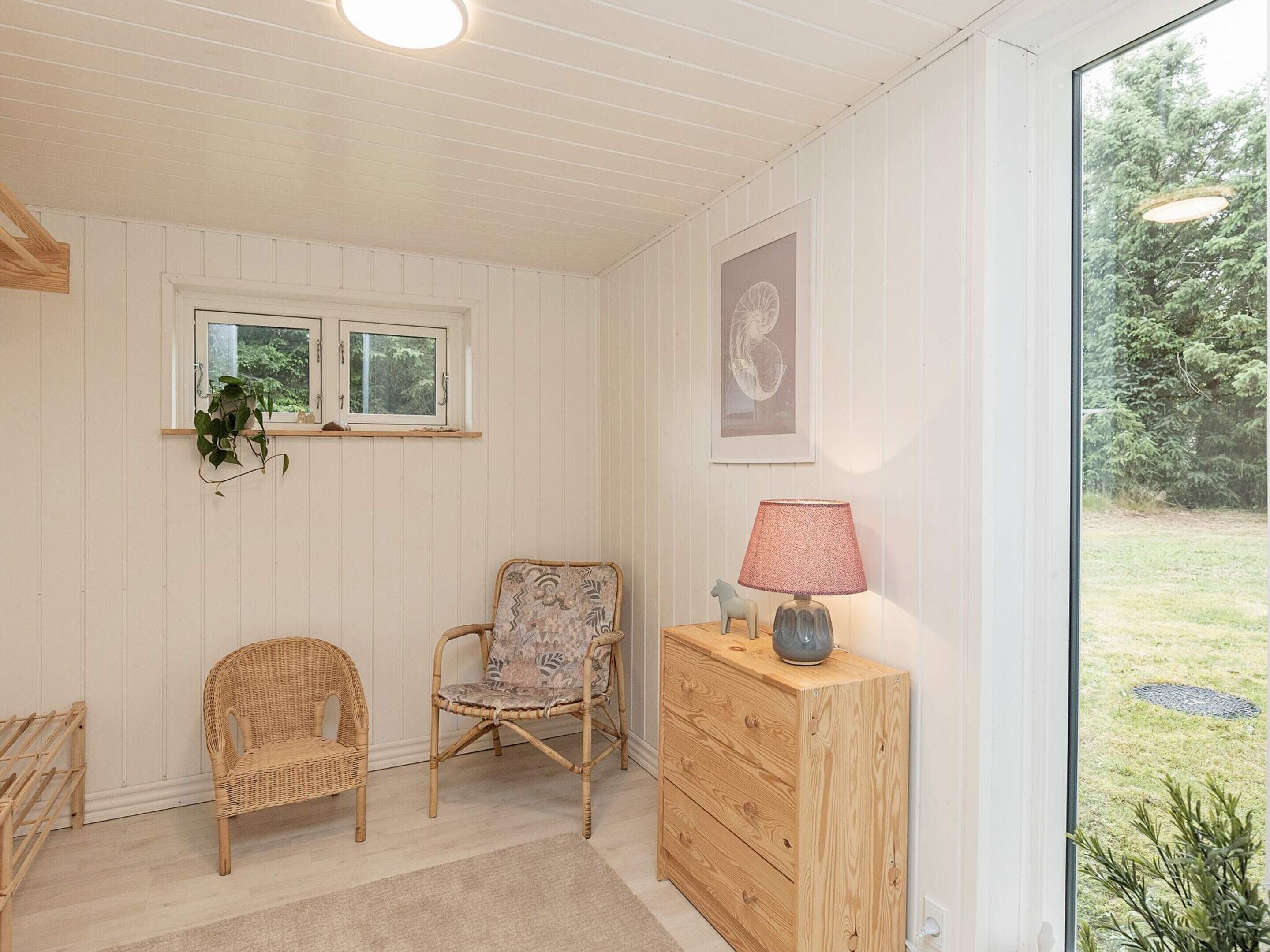 A room with two wicker chairs, a wooden dresser, a lamp, and a window with a view of greenery.