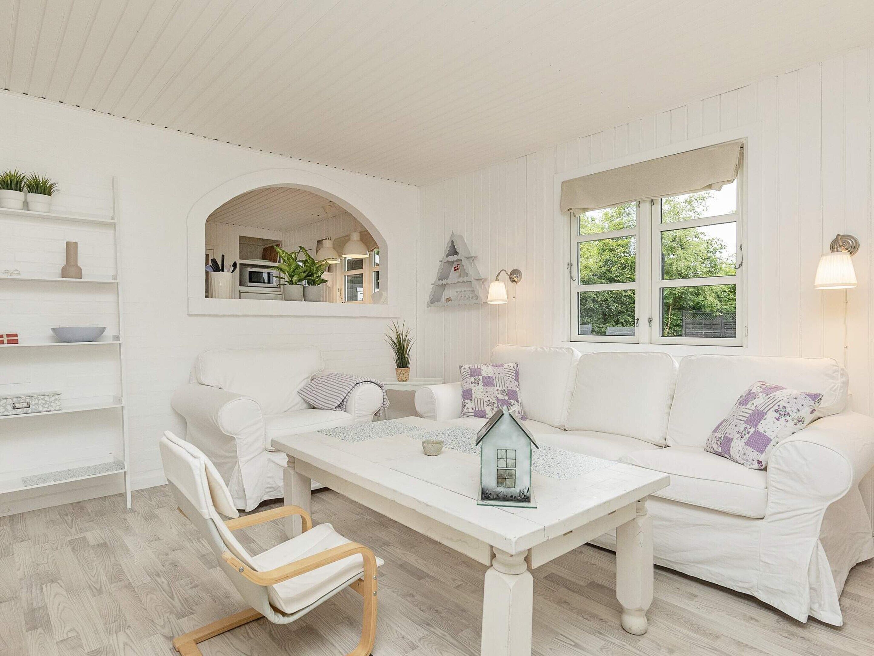 Un intérieur aux couleurs claires, avec un canapé, une table et une chaise. Une fenêtre offre une vue sur la verdure à l’extérieur.