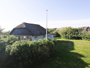 A thatched-roof house with a white exterior, surrounded by lush greenery and a clear sky.