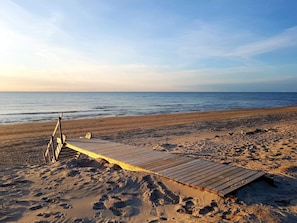 Una playa con un paseo marítimo de madera que conduce al agua, huellas en la arena y un cielo despejado.