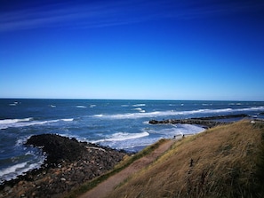 A coastal landscape with a rocky shoreline, waves crashing onto the rocks, and a clear blue sky. - 4 Person Holiday Home in Hjorring (Hjørring)