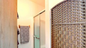 Casa de banho com cabine de duche em vidro, separador de privacidade trançado e toalhas estampadas na parede.