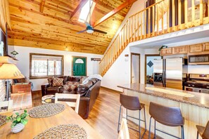 A spacious living area with a high ceiling, wooden beams, a green door, a brown leather sofa, a wooden dining table, and a kitchen with stainless steel appliances. - Near Hiking Trails: Wooded Cabin in Lakeside! (Lakeside)