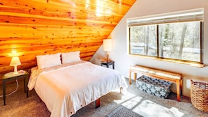 Um quarto aconchegante com teto inclinado, uma cama grande, uma mesa de cabeceira com abajur, um banco e uma janela com vista para árvores.