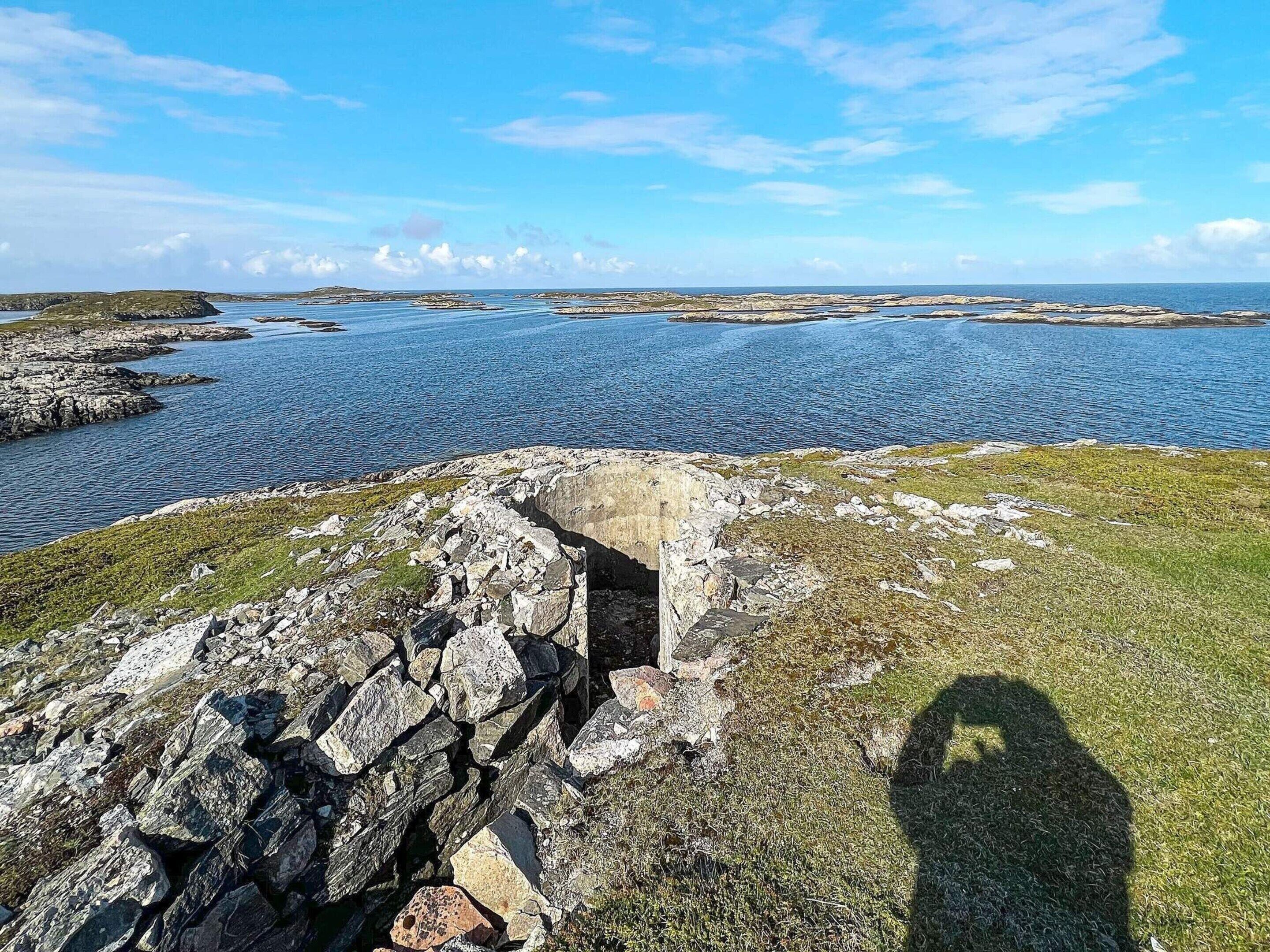 A coastal landscape with a rocky shoreline, a small cave-like structure, and a clear view of the sea.