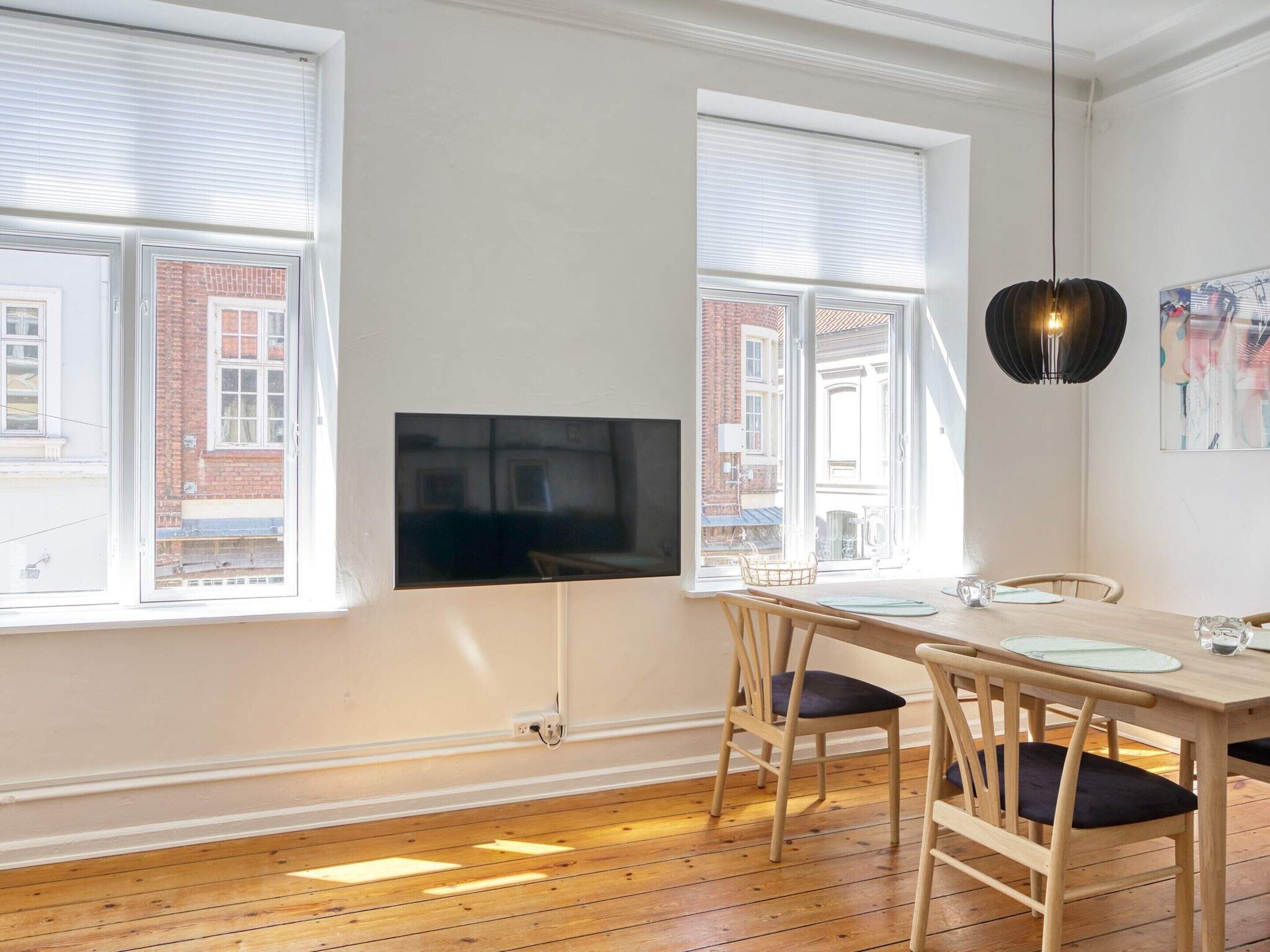 A modern living room with a flat-screen TV, wooden dining table, chairs, and large windows with blinds.