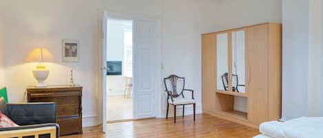 A room with a bed, a wooden wardrobe, a chair, a desk with a lamp, and a door leading to an outdoor area.