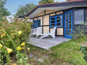 Un bâtiment doté d’une terrasse en bois, de deux fauteuils de jardin blancs et d’une porte bleue.