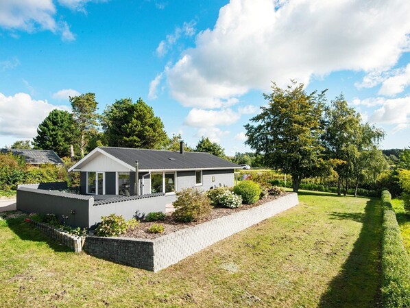 Ett modernt hus med en grå fasad, en inglasad veranda och en välskött trädgård.