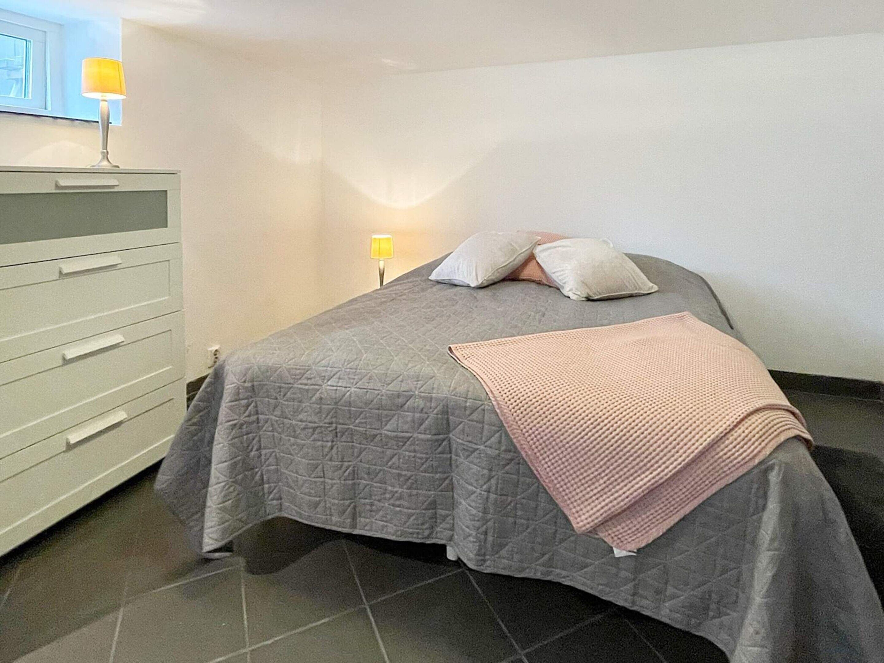 A neatly made bed with a grey quilt and a pink blanket, a white dresser with drawers, and a nightstand with a lamp.