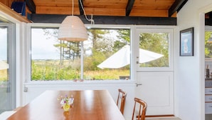 Una mesa de madera con sillas, una lámpara colgante y vistas al exterior a través de amplios ventanales.