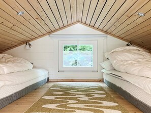 A small attic bedroom with two beds, a window with a view of greenery, and a wooden ceiling.