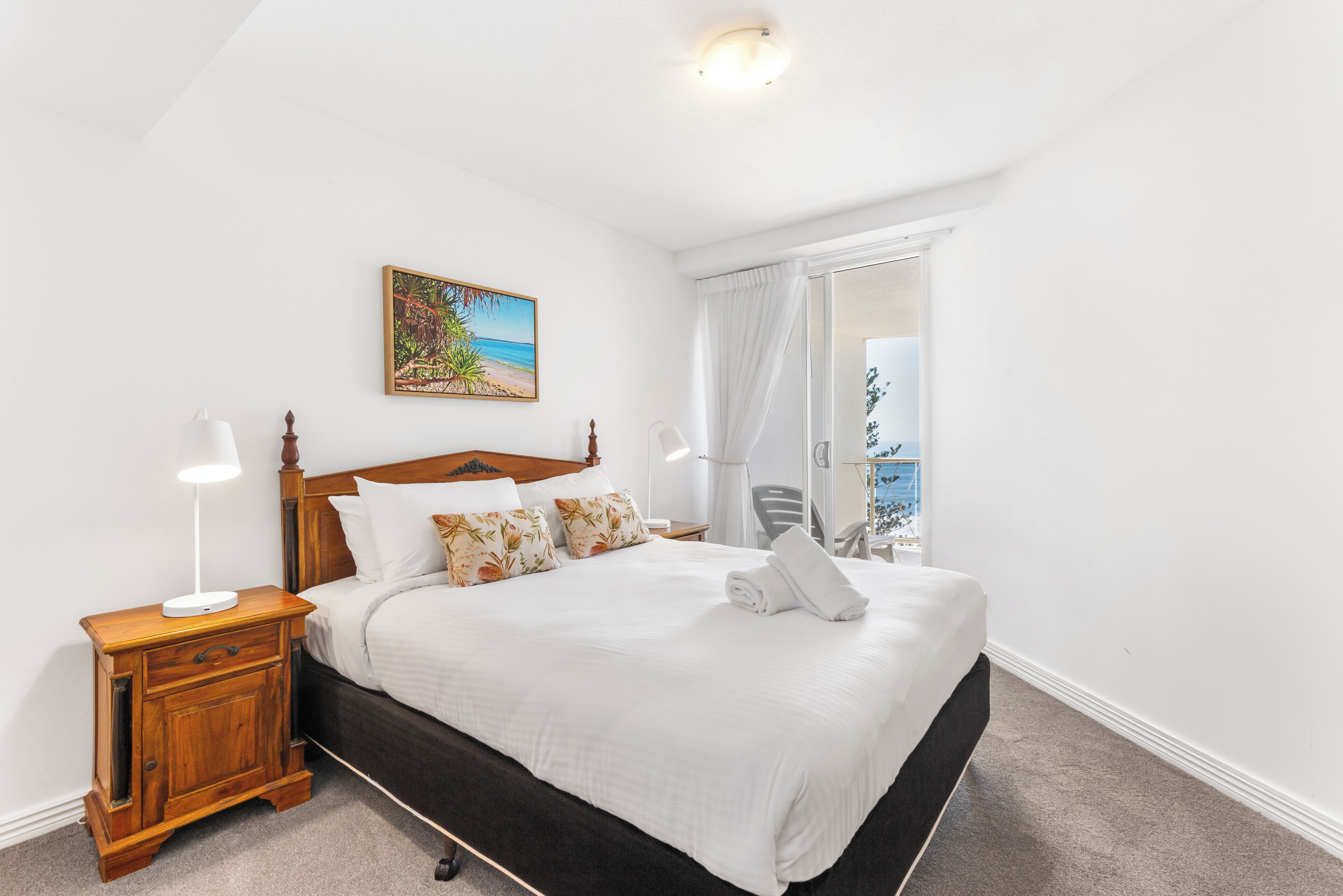 A neatly made bed with white linens and a wooden headboard, a small wooden nightstand with a lamp, and a painting of a beach scene on the wall.
