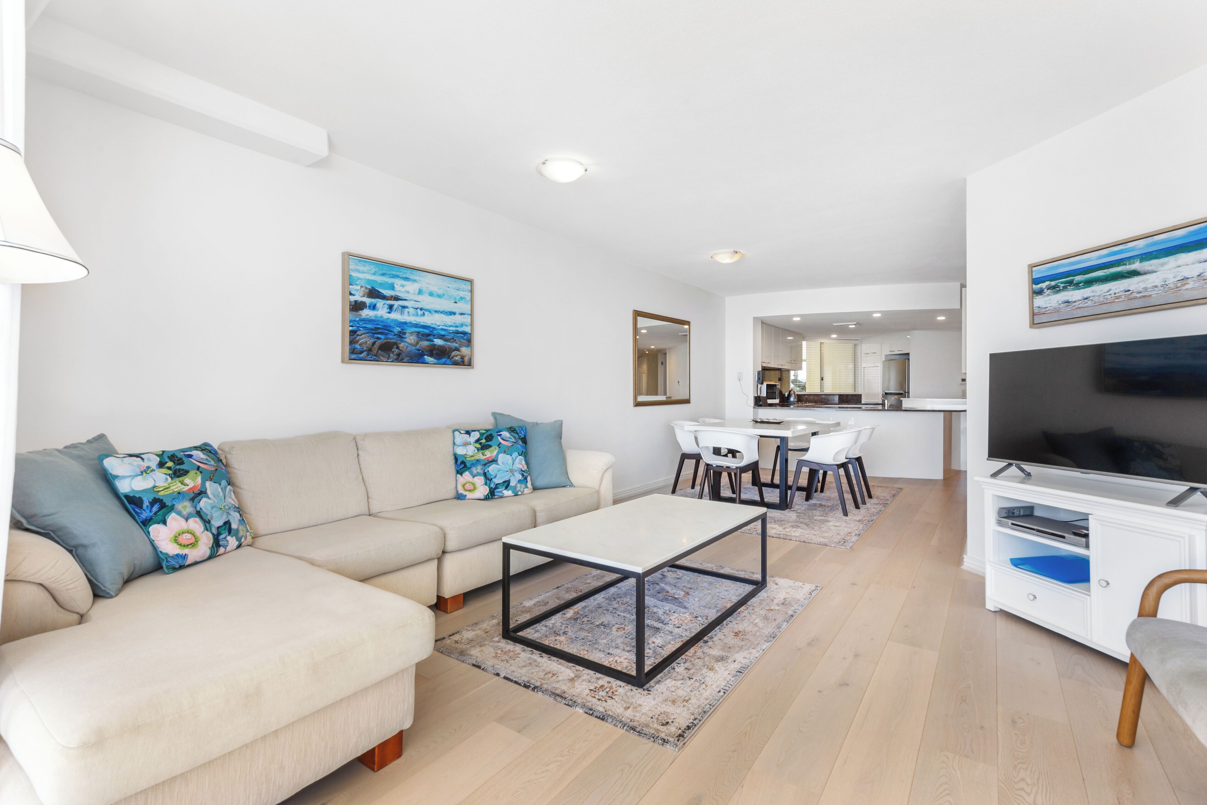 Una sala moderna con sofá, mesa de centro y mueble para televisión. Hay un comedor con sillas y una mesa. Las paredes son blancas y el piso es de madera.