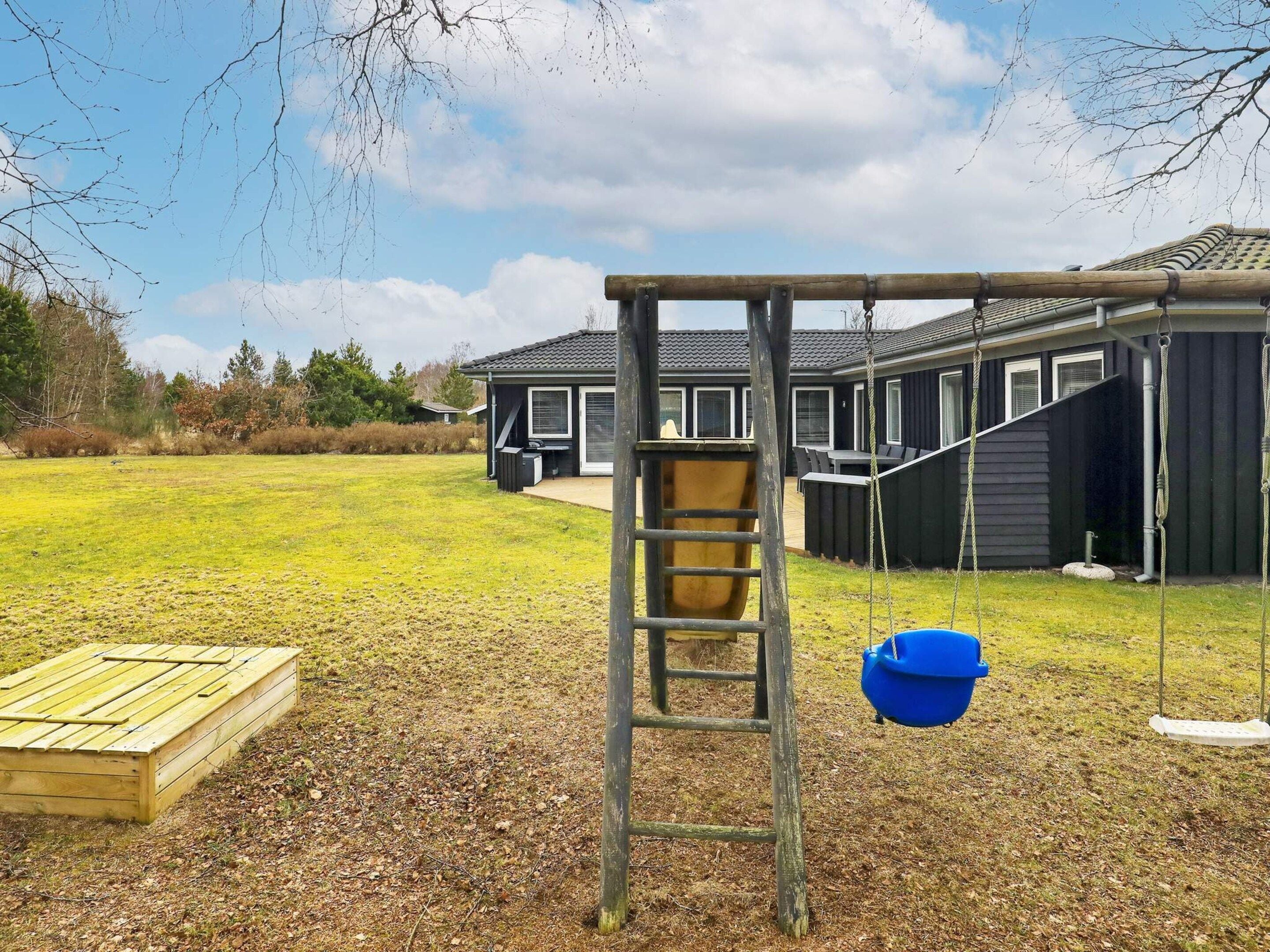 A playground with a swing set and a slide, a wooden play structure, and a house in the background.