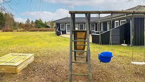 A playground with a swing set and a slide, a wooden play structure, and a house in the background. - 8 Person Holiday Home in Hals (Hals)