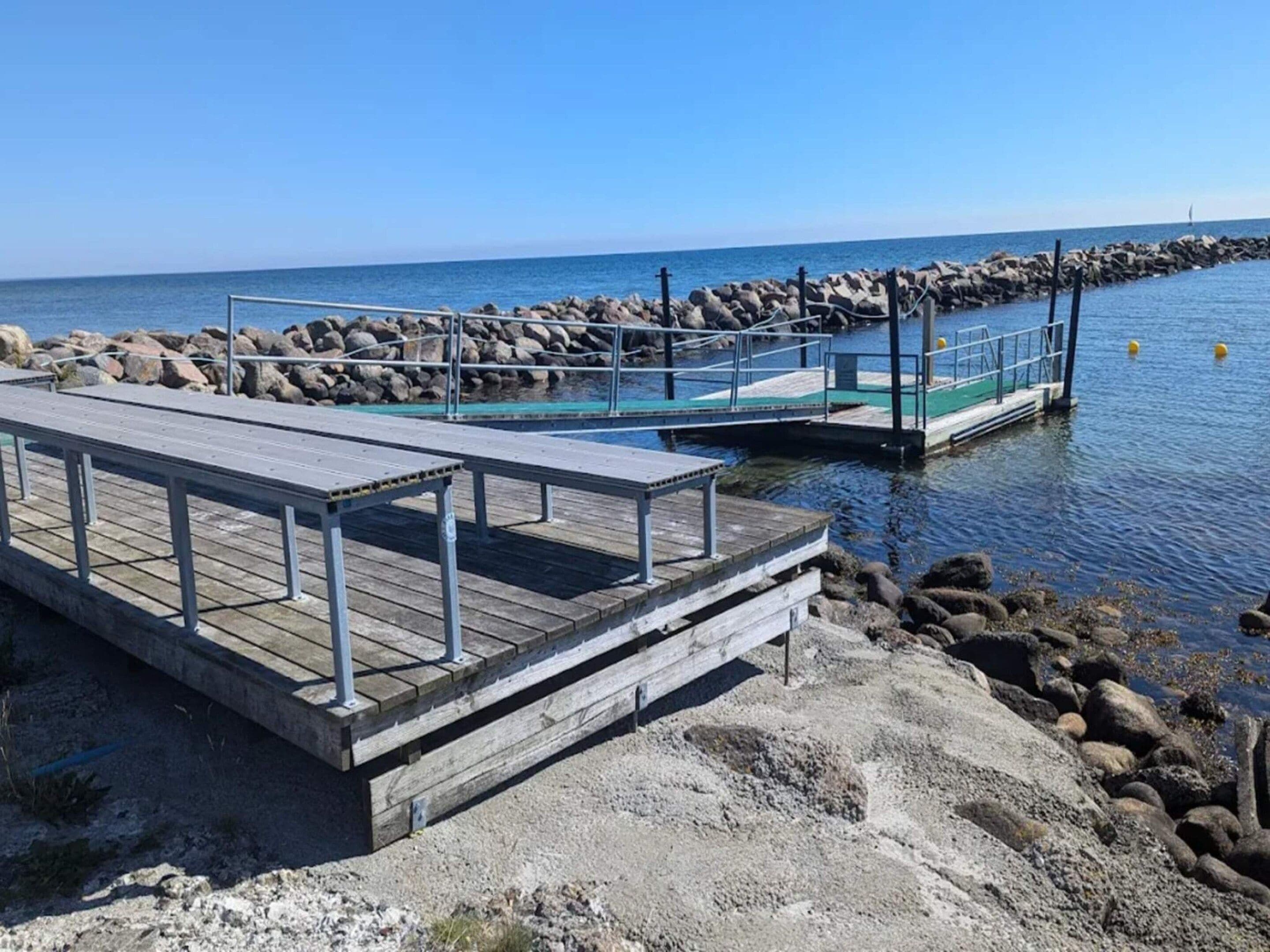 A wooden pier extending into calm waters, with a rocky shoreline and a clear sky.