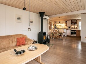 A cozy living room with a wooden ceiling, a sofa with a red cushion, a glass coffee table with remote controls, and a black wood stove.